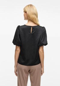 Blusa nera in satin con maniche corte a palloncino e dettaglio a chiave sulla schiena. Indossata con pantaloni beige su misura. Tessuto morbido.