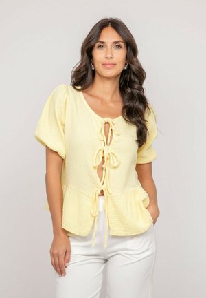 Femme aux longs cheveux ondulés et foncés, portant un chemisier jaune texturé à manches courtes avec des fermetures à nouer à l'avant et un pantalon blanc, sur fond clair.