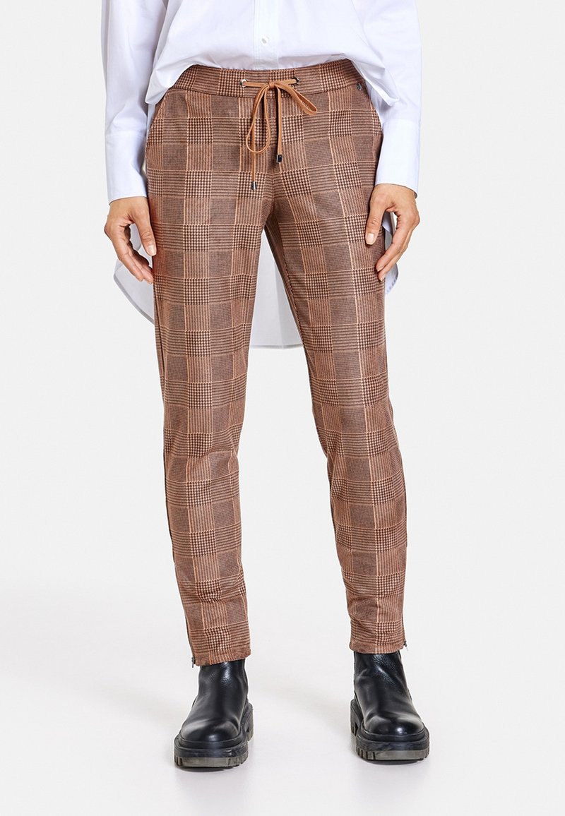 Pantalon en tartan marron avec une taille à cordon, une jambe fuselée et des poignets zippés, associé à des bottines noires. Texture de tissu lisse.