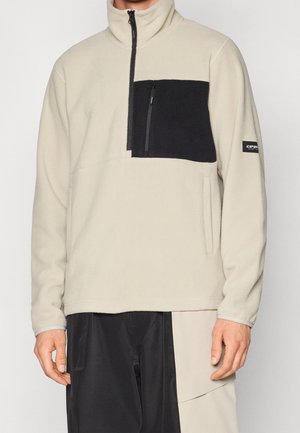 Mann trägt beigen Fleece-Pullover mit schwarzer Brusttasche und Reißverschluss, kombiniert mit zweifarbigen schwarzen und beigen Hosen.