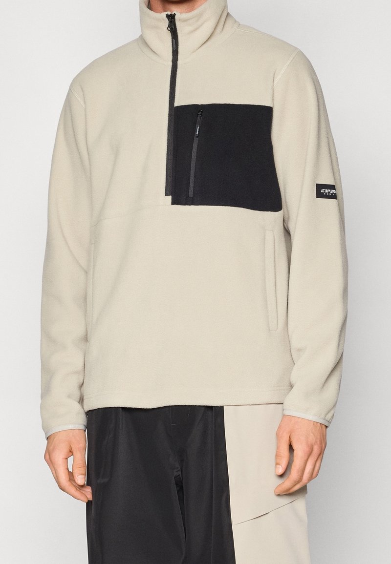 Homme portant un pull polaire beige avec une poche poitrine noire et une fermeture éclair, assorti à un pantalon bicolore noir et beige.