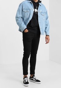 Chaqueta de mezclilla azul clara sobre una sudadera negra con capucha, pantalones negros ajustados y zapatillas negras con detalles blancos. Outfit casual en capas.