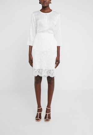 Femme portant un chemisier blanc à manches trois-quarts, une jupe en dentelle blanche et des sandales à talons hauts beiges à brides, debout devant un fond uni.