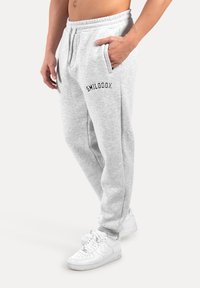Graue Jogginghose mit einem Kordelzug, seitlichen Taschen und schwarzem Logo "SMILODDOX" am linken Oberschenkel. Kombiniert mit weißen Sneakers.