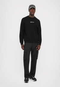 Svart hettegenser med en liten logo, kombinert med svarte cargo-bukser med sidelommer og svarte sneakers med hvite detaljer. Grå caps.