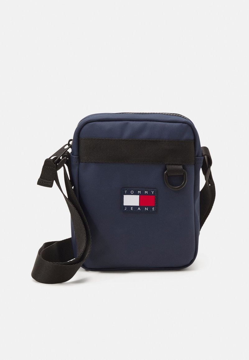 Bolso bandolera de nailon azul marino con una correa negra y un parche rectangular con un logotipo que presenta colores rojo, blanco y azul. Cierre de cremallera.