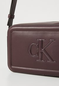 Sac bandoulière Calvin Klein en cuir bourgogne foncé, présentant un logo en relief proéminent, une texture lisse et des bords cousus.