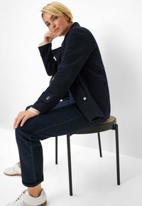 Marineblauw wollen blazer in een wolmix met een gestructureerde afwerking, voorzien van een knoopmanchet, gecombineerd met donkere denim jeans en witte sneakers.