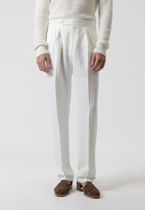 Hombre con pantalones blancos plisados, suéter de manga larga texturizado color blanco roto y sandalias cerradas de cuero marrón, de pie frente a un fondo liso.