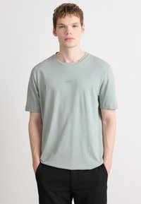 Light green cotton t-shirt with short sleeves, round neckline, and subtle embossed logo on the chest. Paired with black trousers.