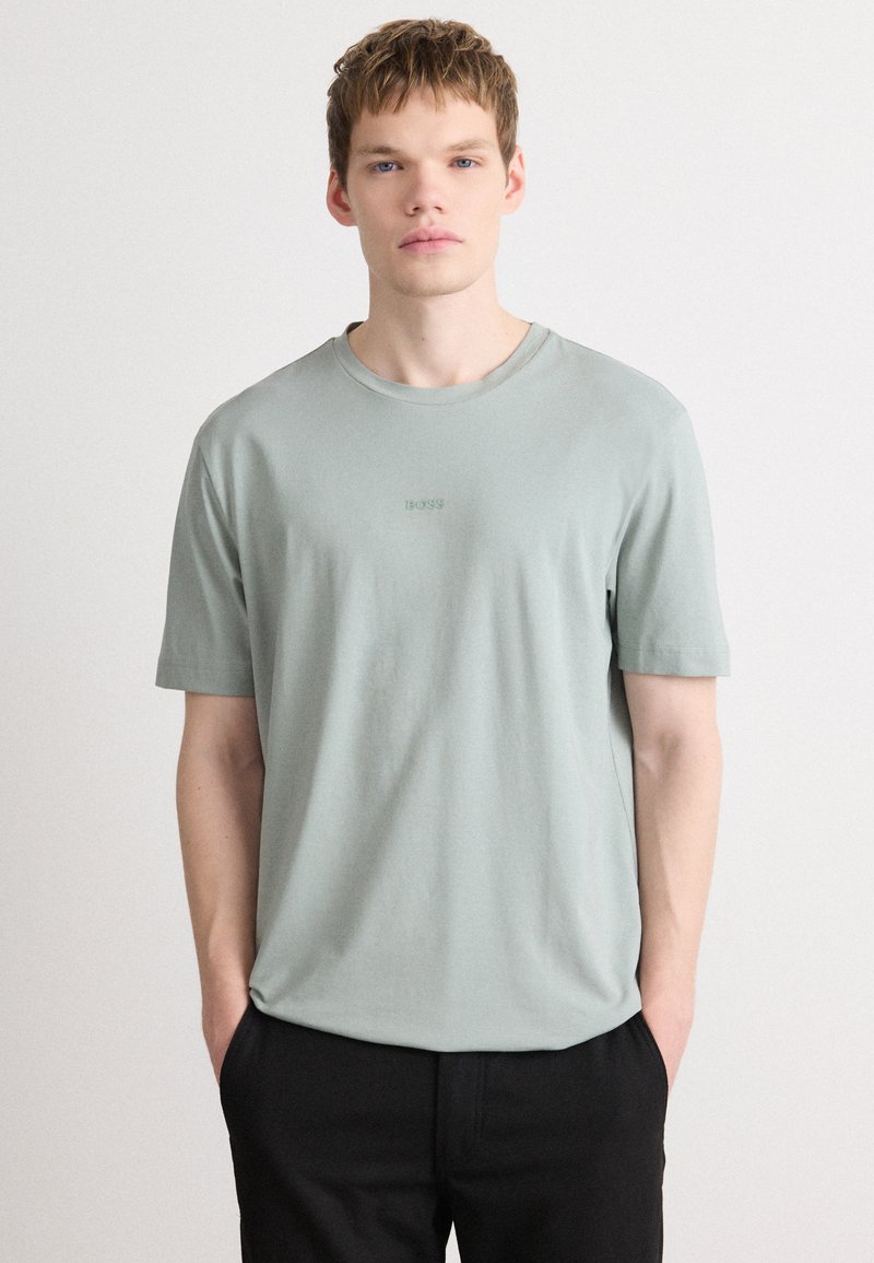 Light green cotton t-shirt with short sleeves, round neckline, and subtle embossed logo on the chest. Paired with black trousers.