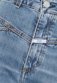 Jean en denim bleu avec des coutures orange, une poche avant et une étiquette avec "CLOSED" visible sur la taille.