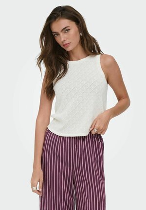 Kvinde iført en ærmeløs hvid tekstureret top og højtaljede lilla bukser med vertikale hvide striber, poserer foran en ensfarvet baggrund.