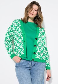 Cardigan verde con motivo a cuori bianchi, chiusura a bottoni e polsini a costine, abbinato a una maglietta verde uni e jeans blu.