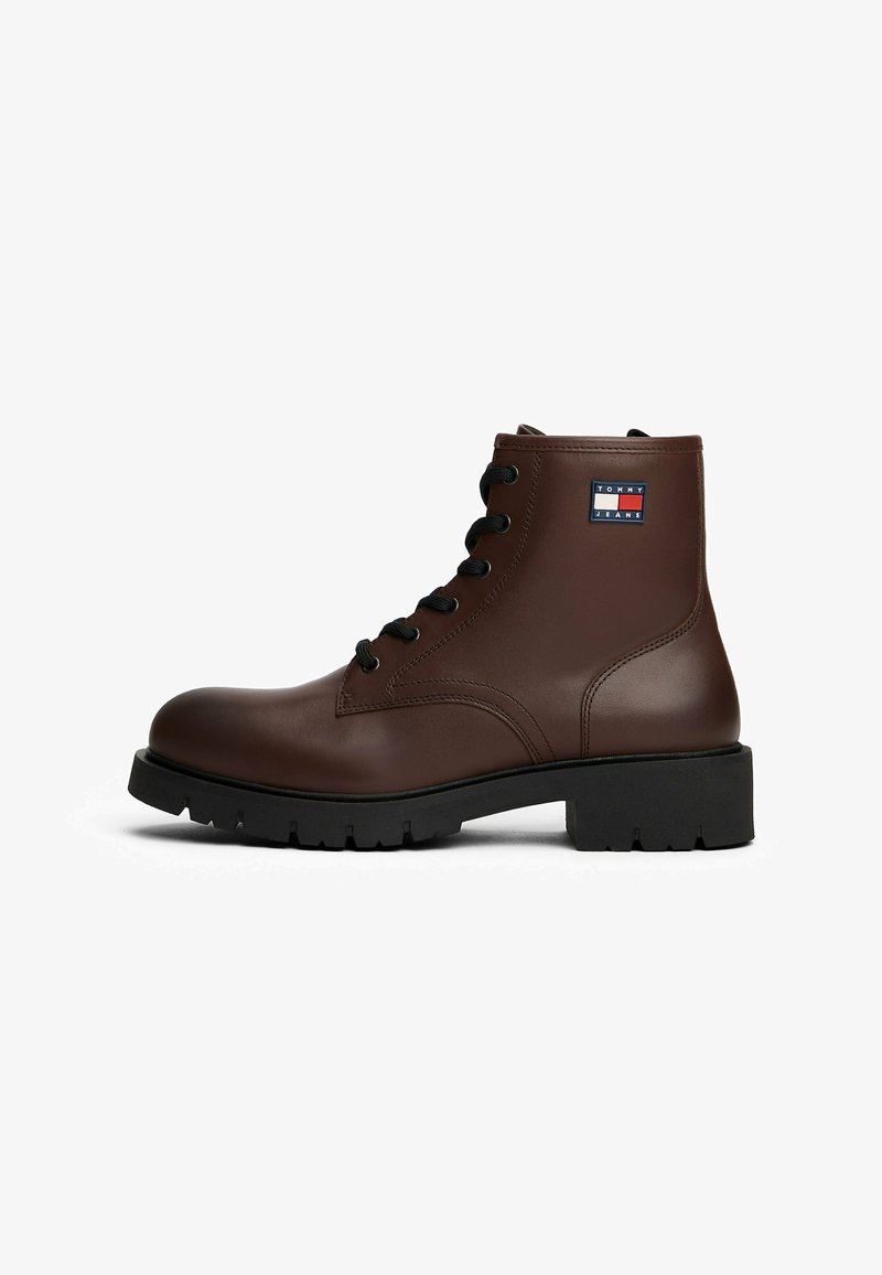Braune Lederstiefeletten mit schwarzen Schnürsenkeln, ausgestattet mit einer dicken schwarzen Gummisohle und einem kleinen Logopatch an der Außenseite.