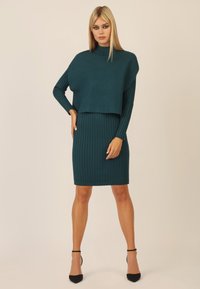 Tee-grünes Strickkleid mit geripptem Muster, hohem Kragen und schmal zulaufenden Ärmeln. Der Rock ist ebenfalls gerippt, kniefest und wird mit schwarzen, hohen Schuhen kombiniert.
