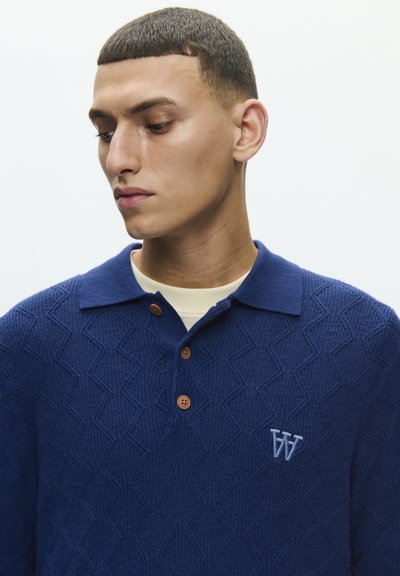 Polo bleu marine texturé avec un motif en losange embossé, doté de boutons marron et d'un logo brodé sur la poitrine.