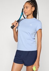 Ανοιχτό μπλε κοντομάνικο μπλουζάκι με στρογγυλή λαιμόκοψη και λευκό λογότυπο, σε συνδυασμό με σκούρο μπλε σορτς, κρατώντας ρακέτα και μπάλα του τένις.