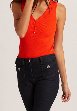 Vrouw draagt een mouwloze oranje geribde top met knopen, gecombineerd met hoog getailleerde zwarte spijkerbroek met decoratieve voorzakken.