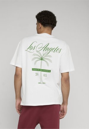 Mann, der von hinten zu sehen ist, trägt ein weißes T-Shirt mit grünem Palmenmotiv und dem Schriftzug „Los Angeles Urban Classics 2005“, kombiniert mit dunkelroter Hose.