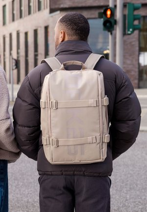 Férfi fekete steppelt dzsekiben és bézs hátizsákkal, aki egy városi utcán áll egy piros lámpás gyalogátkelő mellett.