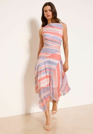 Femme portant une robe midi sans manches à rayures pastel rose, violettes et blanches avec un ourlet asymétrique, marchant en talons beige à bride à la cheville.