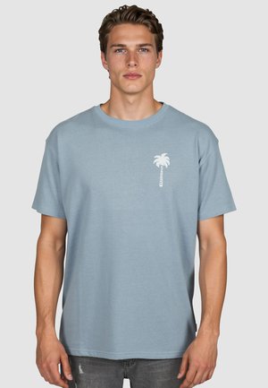 Junger Mann mit kurzen braunen Haaren, der ein hellblaues T-Shirt mit einer kleinen weißen Palmen-Grafik auf der linken Brust trägt.