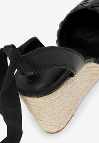Espadrille à talon compensé en cuir noir avec semelle en jute tressé. Présente une large bride et une surface texturée. Bride de cheville en tissu noir ajustable.