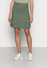 Žena v zelené, vzorované sukně ke kolenům, světle zeleném topu a bílých teniskách stojící na jednobarevném pozadí.