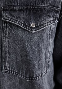 Tasca della giacca di jeans nera con patta fissata da un singolo bottone a pressione argentato e cuciture visibili su tessuto testurizzato.
