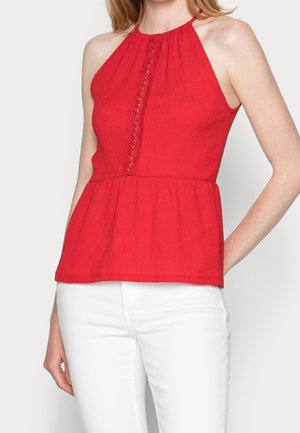 Woman wearing sleeveless red halter top with lace detail down front paired with white fitted pants, hand resting on hip.