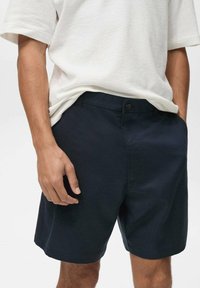 Shorts bleumarin dintr-un material ușor, cu un croi drept, închidere cu nasture și buzunare laterale; asortați cu un tricou de culoare deschisă.
