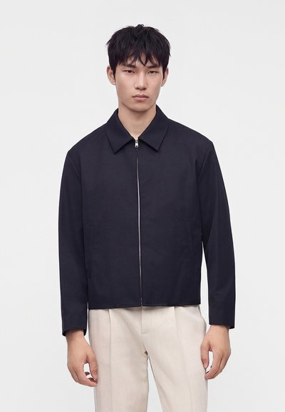 Jeune homme aux cheveux courts et foncés, portant une veste noire zippée et un pantalon beige, debout devant un fond clair uni.