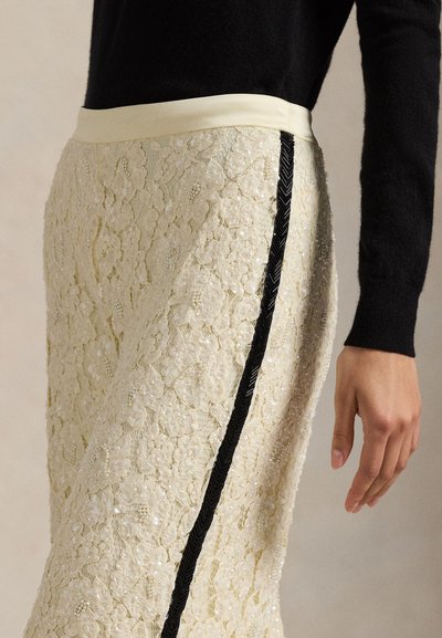 Spetskjol i krämfärg med intrikata blommönster, prydd med pärlor; har en kontrasterande svart sidopanel och ett krämfärgat midjeband.