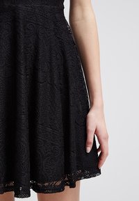 Schwarzes Spitzenkleid mit einem ausgestellten Rock, der aufwendige wirbelnde Muster und einen zarten Saum aufweist. Nahaufnahme der Stofftextur und Handplatzierung.