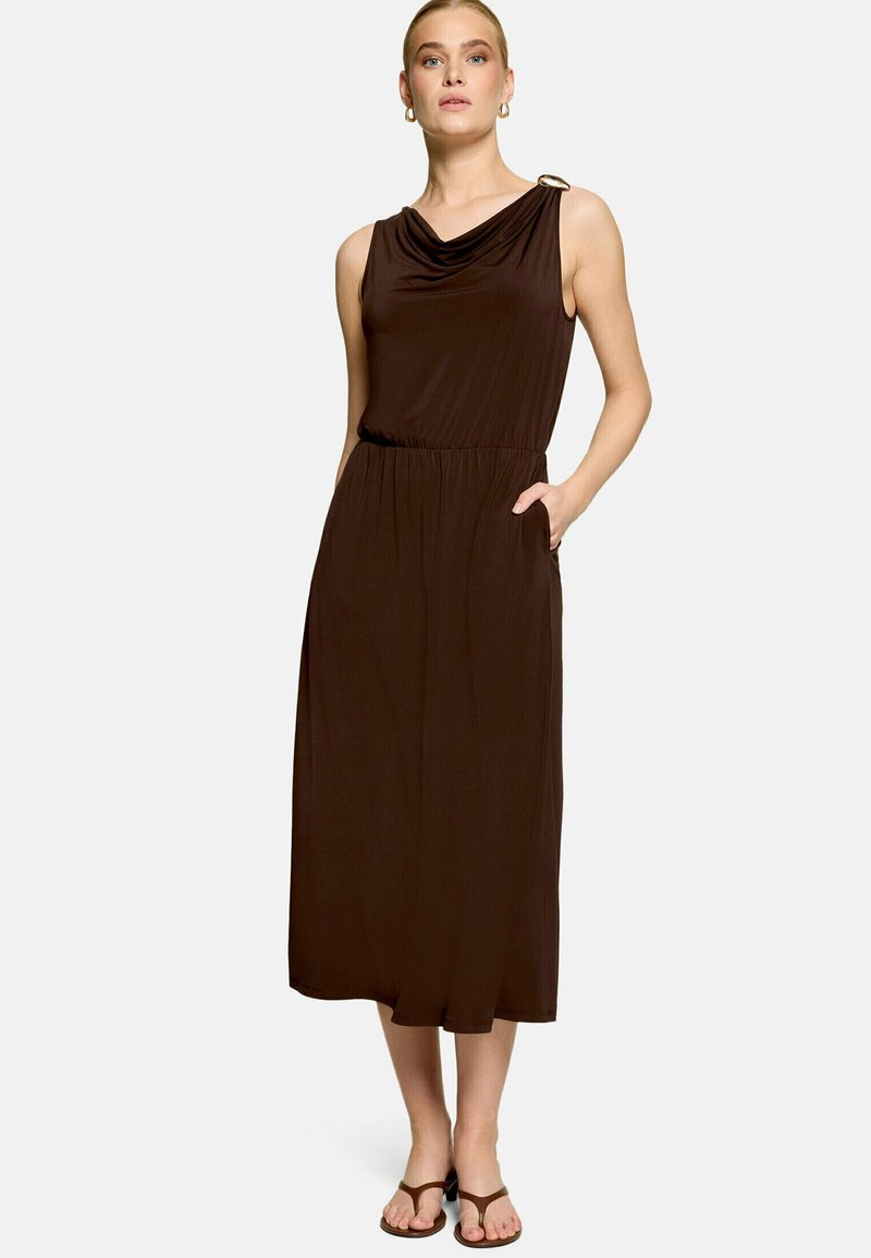 Femme portant une robe midi sans manches marron foncé avec un col drapé, des poches latérales et des sandales marron, debout avec une main dans la poche.