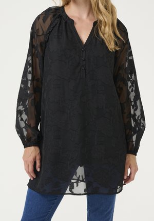 Femme portant un chemisier noir transparent à manches longues avec motif floral, boutons et poignets élastiques, associé à un jean bleu.