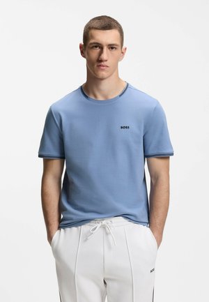 Jeune homme aux cheveux courts portant un t-shirt BOSS bleu clair et un pantalon blanc à cordon, debout les mains dans les poches devant un fond blanc.