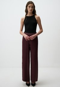 Zwart aansluitend tanktop gecombineerd met een wijde broek met een rood en marineblauw geometrisch patroon. Model staat in zwarte puntige hakken.