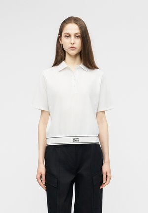 Jauna sieviete ar gariem brūniem matiem, valkājot baltu īsroku polo kreklu ar "JOOP! JEANS" logotipu un melnas kargo bikses uz balta fona.