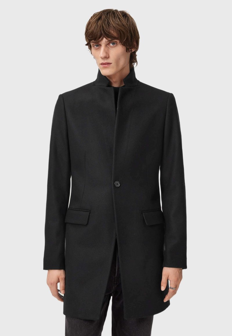 Musta villasekoitteinen takki, jossa on korkea kaulus, yksittäinen napitus, kaksi etutaskua ja suora siluetti. Sujuva rakenne, minimalismi.