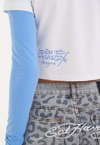 Lyhyt cropped-valkoinen t-paita, jossa on siniset pitkät hihat ja sininen "Don Ed Hardy designs" -teksti. Harmaat farkut, joissa on leopardikuosi ja valkoinen logo.