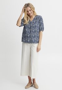 Top blu navy con motivo a onda bianca, design a V, maniche corte; abbinato a pantaloni beige a gamba larga e mocassini chiari.