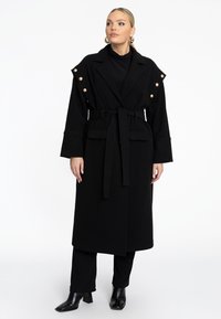Zwarte coat met een brede tailleband, grote revers, decoratieve knopen op de schouders, zijzakken en loszittende mouwen. Draag het over een zwarte broek en laarzen.