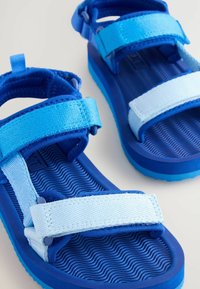 Blå sandaler med två justerbara remmar, tillverkade av texturerat tyg, med ett vågigt mönster på sulan och hållbara gummidetaljer.