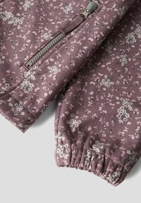 Gros plan sur un tissu mauve à petits motifs floraux blancs, montrant une fermeture éclair argentée et un poignet élastique sur la manche d'un vêtement.