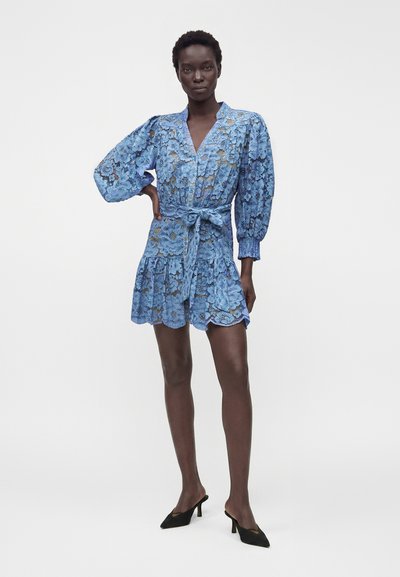 Femme debout portant une robe en dentelle florale bleue avec manches bouffantes, taille ceinturée et mules à talons pointues noires sur fond blanc uni.
