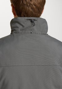 Chaqueta gris con un cuello alto, tejido texturizado y un cordón que se ajusta en la parte posterior. Presenta botones de presión para un cierre seguro.