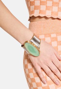Bracciale a larghe manchette con una pietra verde a forma ellittica, circondata da accenti dorati e una banda metallica argentata.