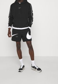 Hombre vestido con una sudadera negra de Nike con logotipos de rayas en las mangas, pantalones cortos negros de Nike con grandes logotipos Swoosh, calcetines blancos y zapatillas negras y blancas de Nike.